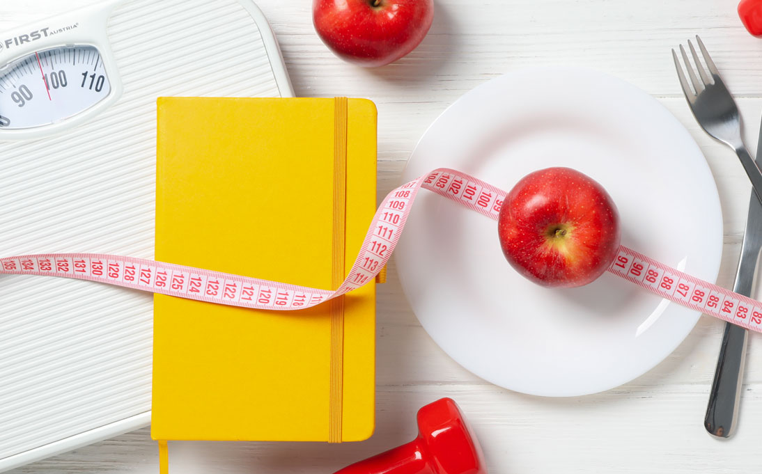 A red apple on top of a red plate, weight scale, and measuring tape lay across a table.