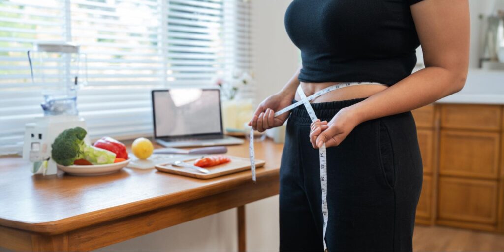 Woman with a measuring tape around her waist next to a table with vegetables on it.