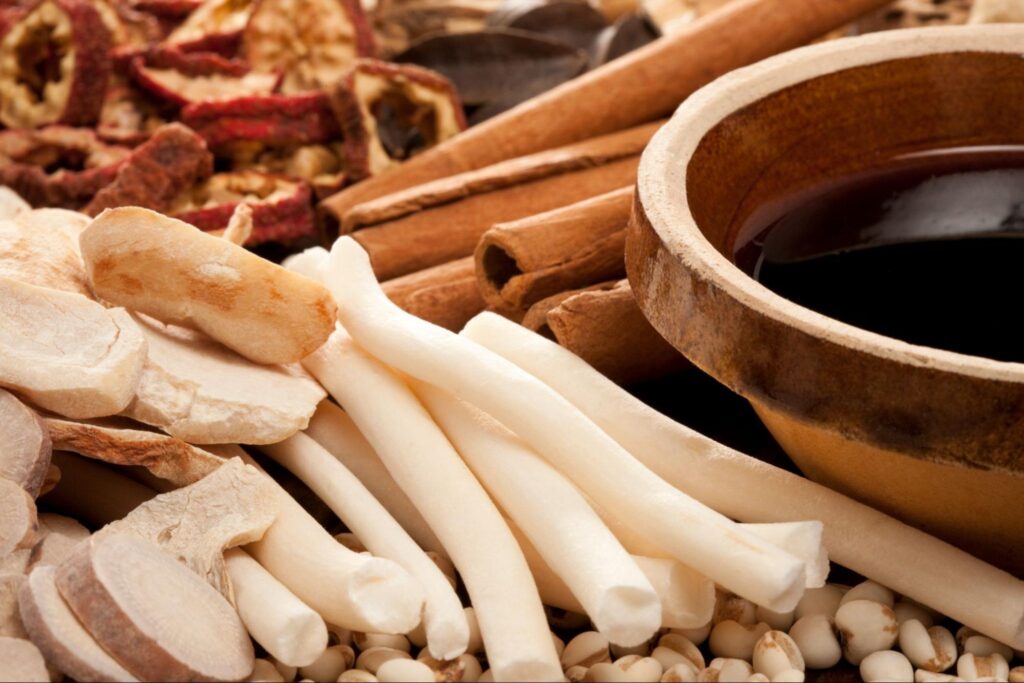 A close-up of various Chinese herbal medicines, including dried herbs and roots, arranged around a bowl of herbal liquid.