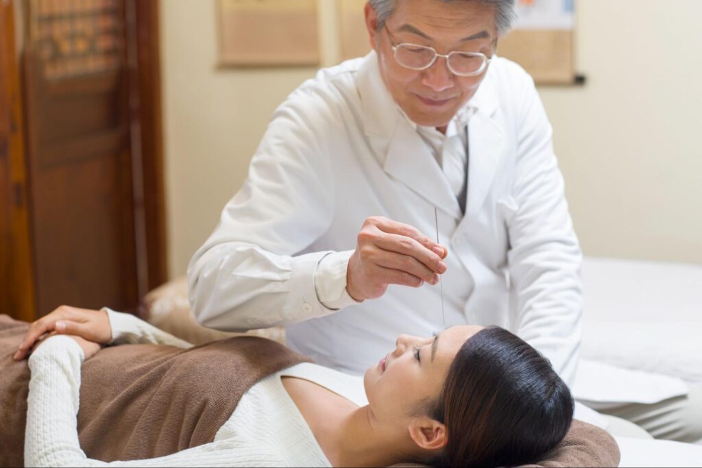 A young woman receives acupuncture on her head from a senior Chinese doctor in a traditional Chinese medical clinic.