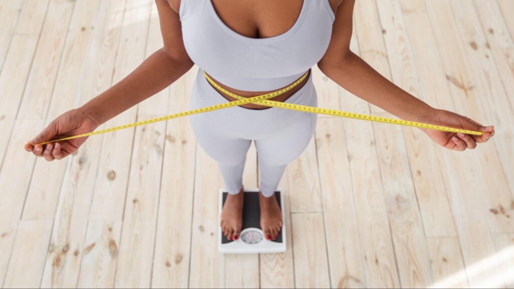 Woman on a scale in a workout set and holding a measuring tape around her waist.